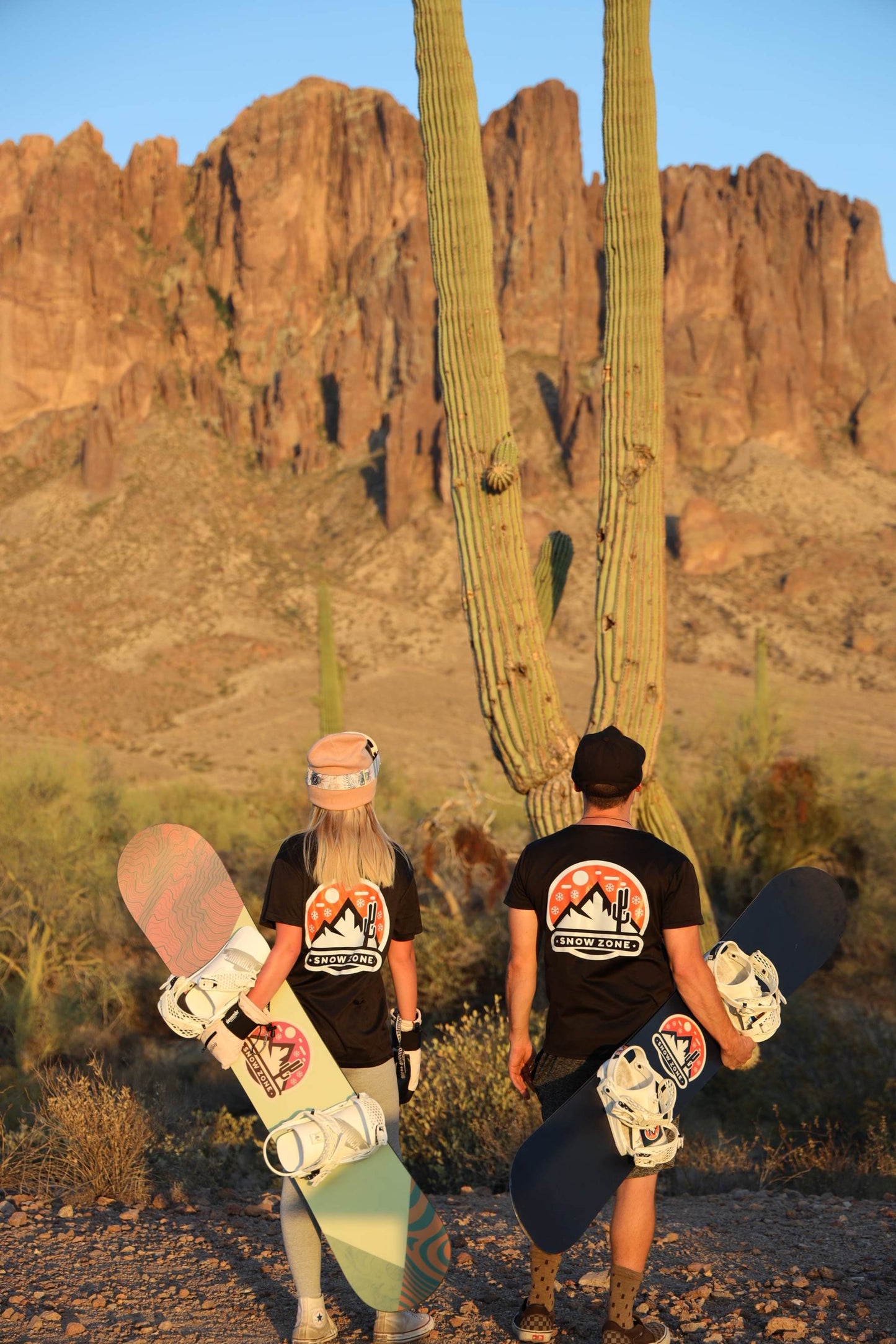 Two people wearing black dry fit short sleeve shirts carrying snowboards in desert landscape with cactus and rocky mountains