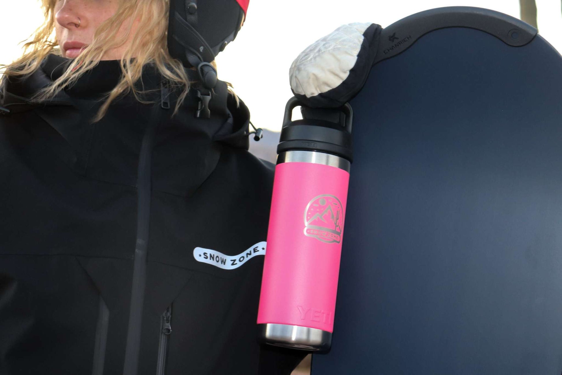 Black insulated snow jacket with pass holder near wrist and zippered pocket, worn by person holding snowboard and pink water bottle.