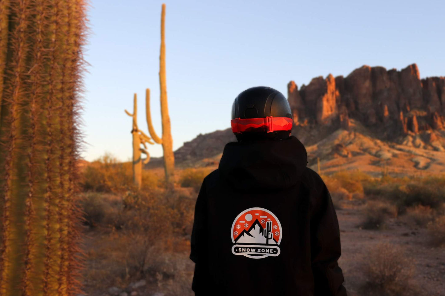 Back view of person wearing insulated snow jacket with hood and helmet in desert landscape with cacti and mountains at sunset
