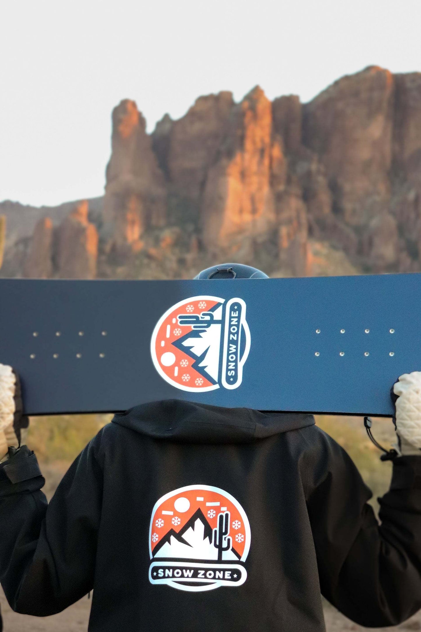 Person wearing insulated snow jacket holding snowboard with snow zone logo outdoors