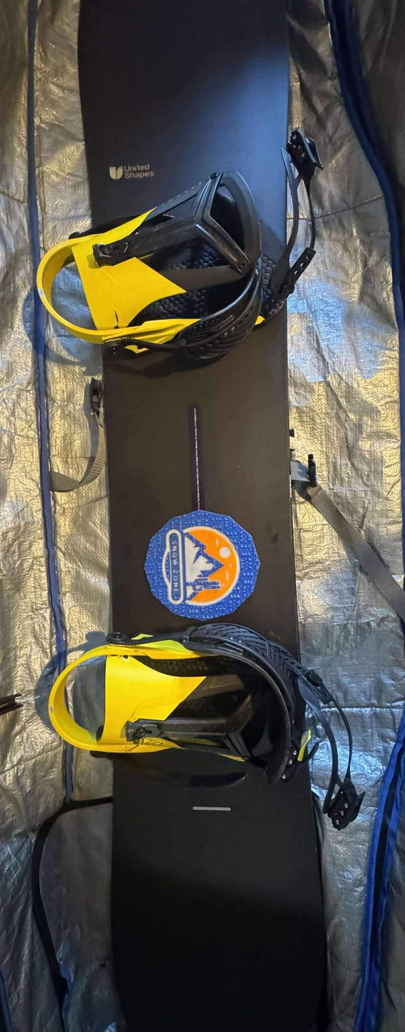 Black snowboard with yellow bindings featuring a Sticky_ride stomp pad and a blue logo patch.