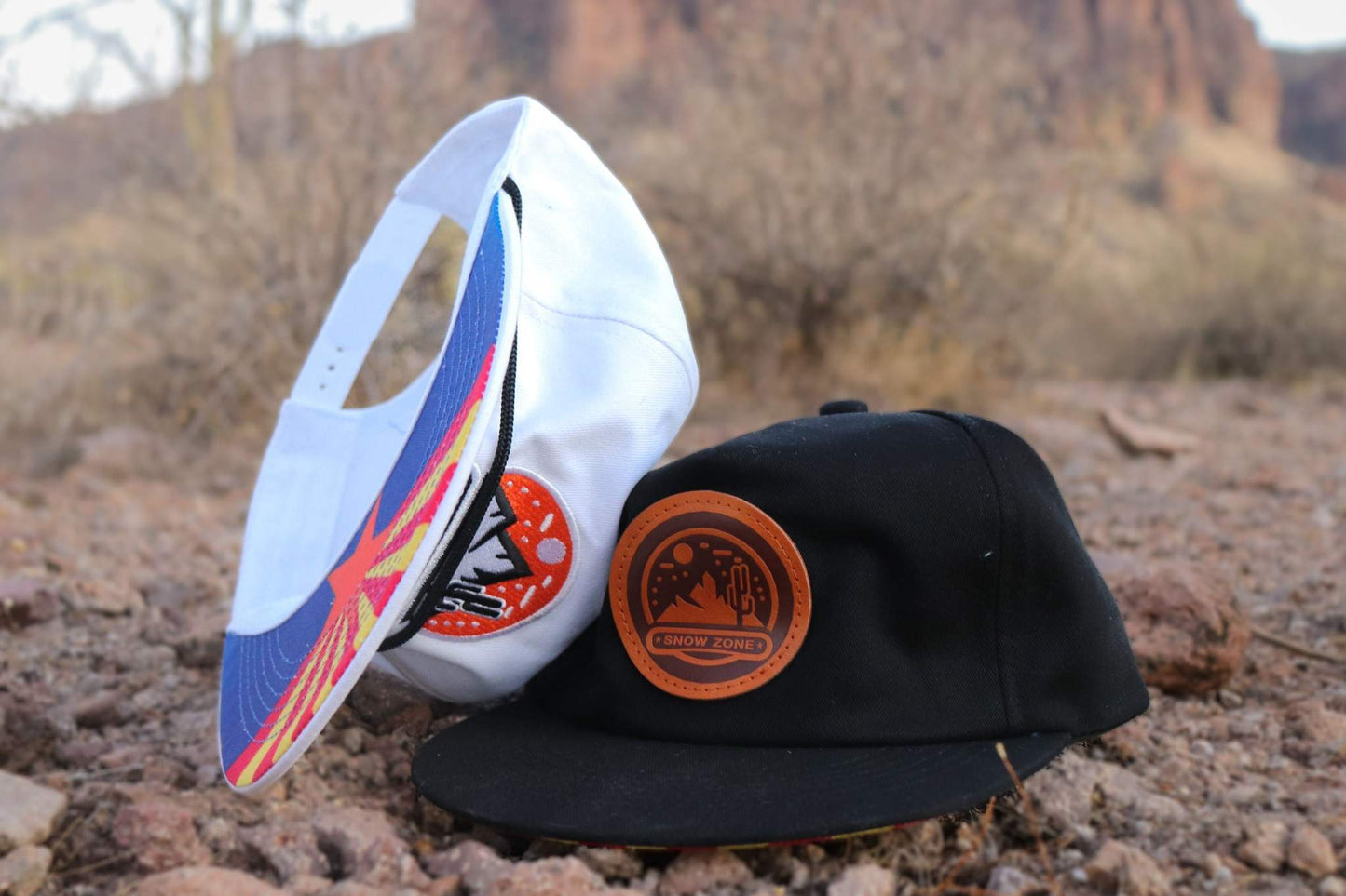 White hat with colorful embroidery patch displayed outdoors on rocky ground next to a black hat
