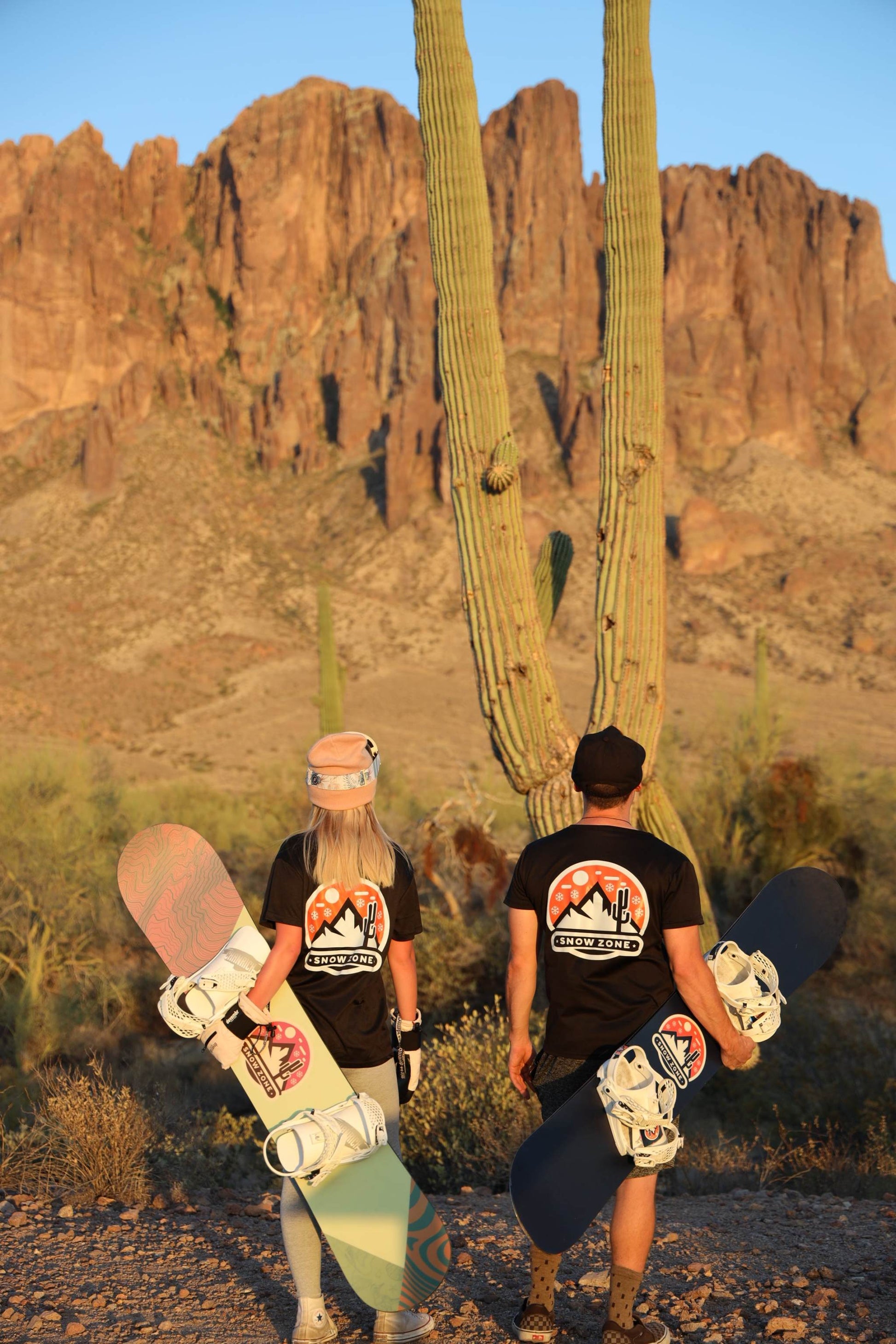Two people wearing black dry fit short sleeve shirts carrying snowboards in desert landscape with cactus and rocky mountains